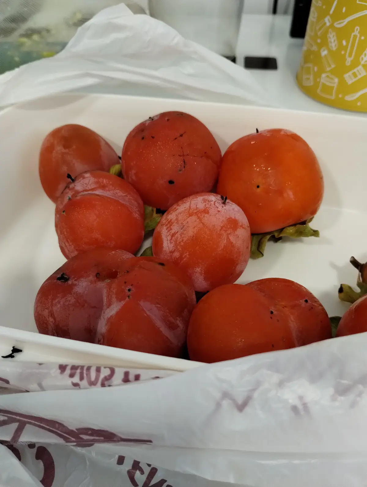 A white container holds several orange persimmons with visible black spots and blemishes, suggesting spoilage. A white…
