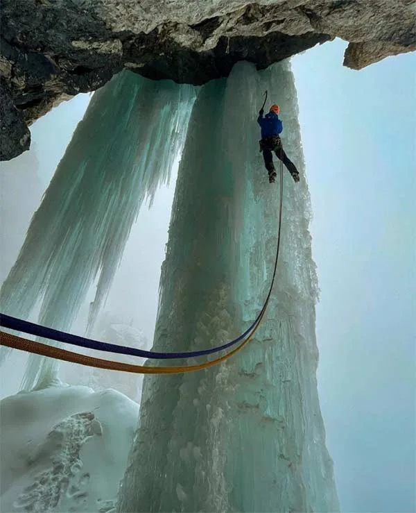 A climber in a blue jacket and orange helmet is rappelling down a massive, translucent ice formation under a rocky overhang…