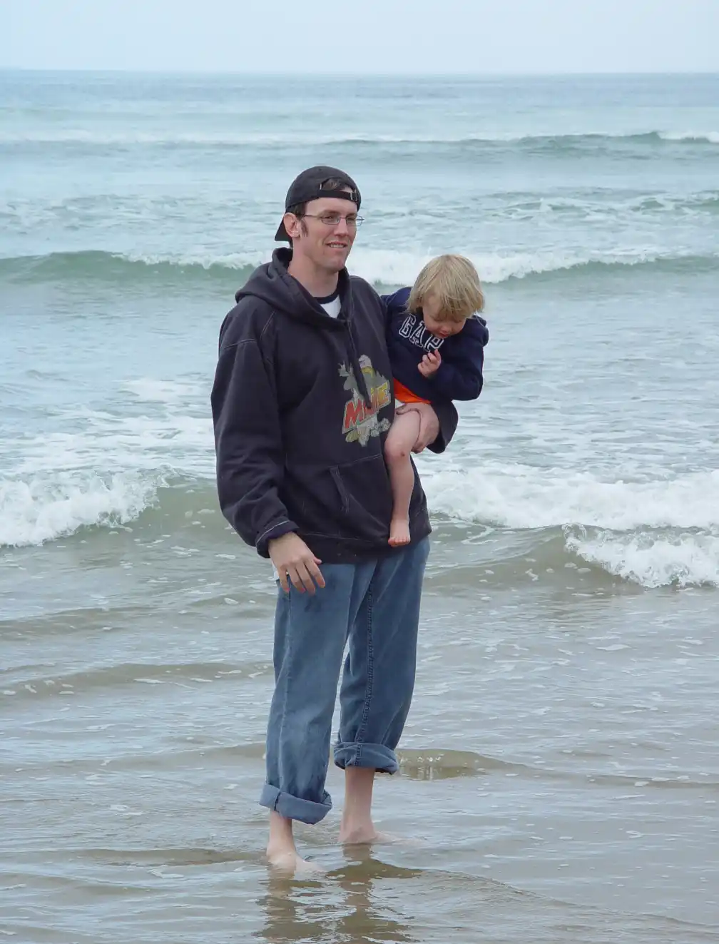 A man wearing a black hoodie and jeans stands barefoot in shallow ocean water, holding a small child in his arms. The child…
