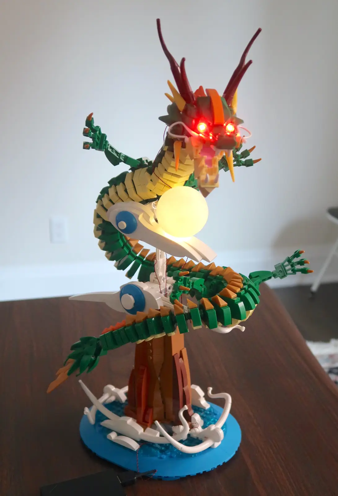 A LEGO dragon model with glowing red eyes and a bright white orb in its mouth, standing on a blue base with white wave…