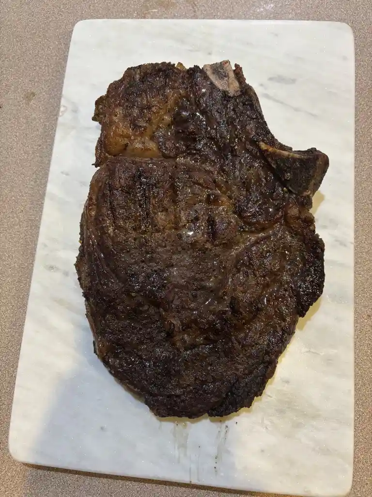 A large, heavily charred, and blackened steak with visible grill marks rests on a white marble cutting board. The meat…