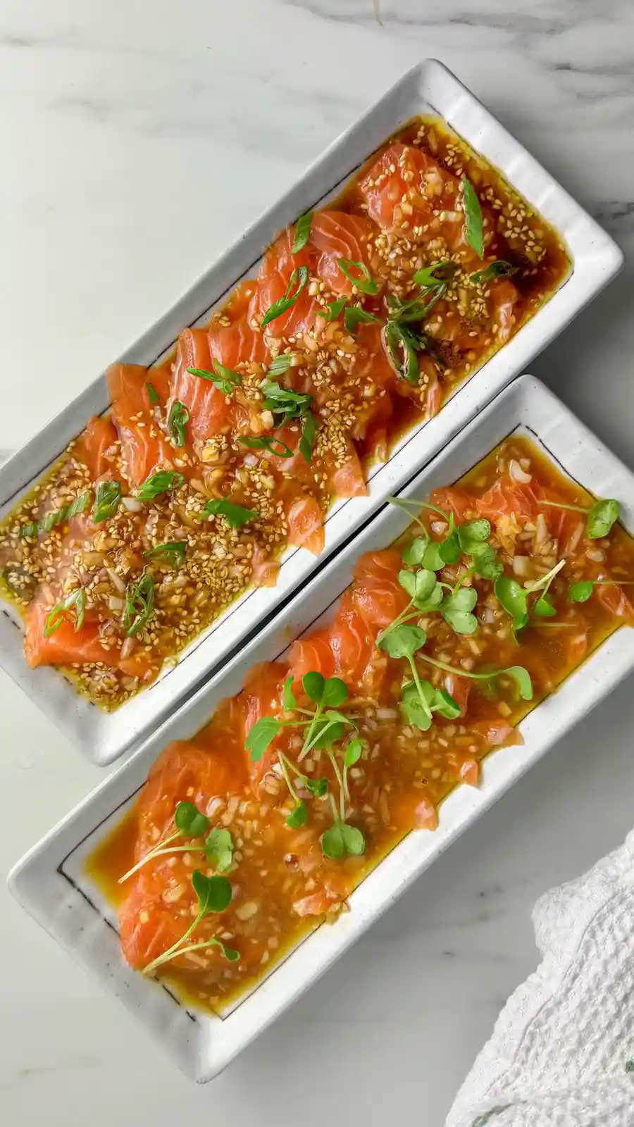 Two white rectangular plates hold raw salmon sashimi slices in a brown sauce, topped with sesame seeds and green garnishes…