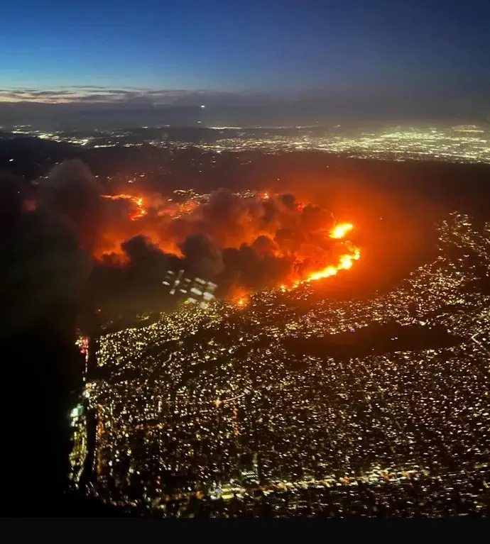 An aerial night photo shows a massive wildfire burning intensely with bright orange flames and thick smoke, engulfing the…