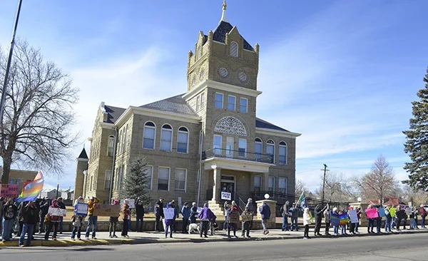 A large group of people stands in a line on a street in front of a large stone courthouse under a clear blue sky. Many hold…