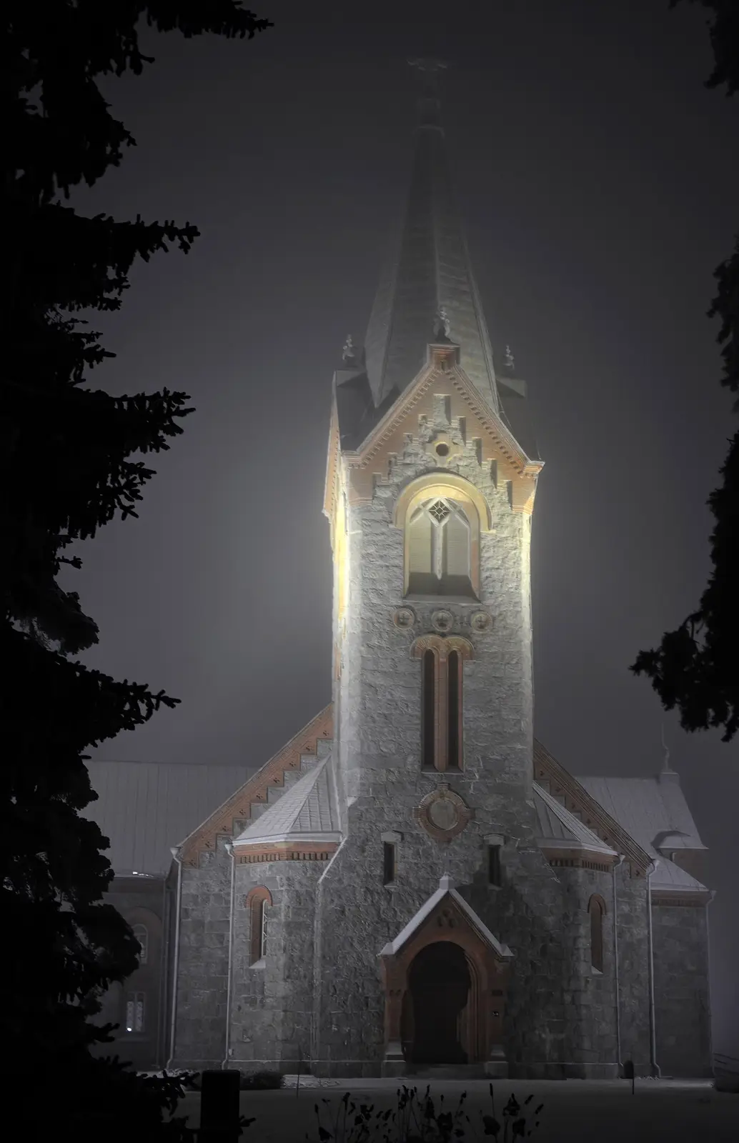 A dark, atmospheric night photo of a stone church with a tall spire, illuminated by artificial light against a foggy sky…