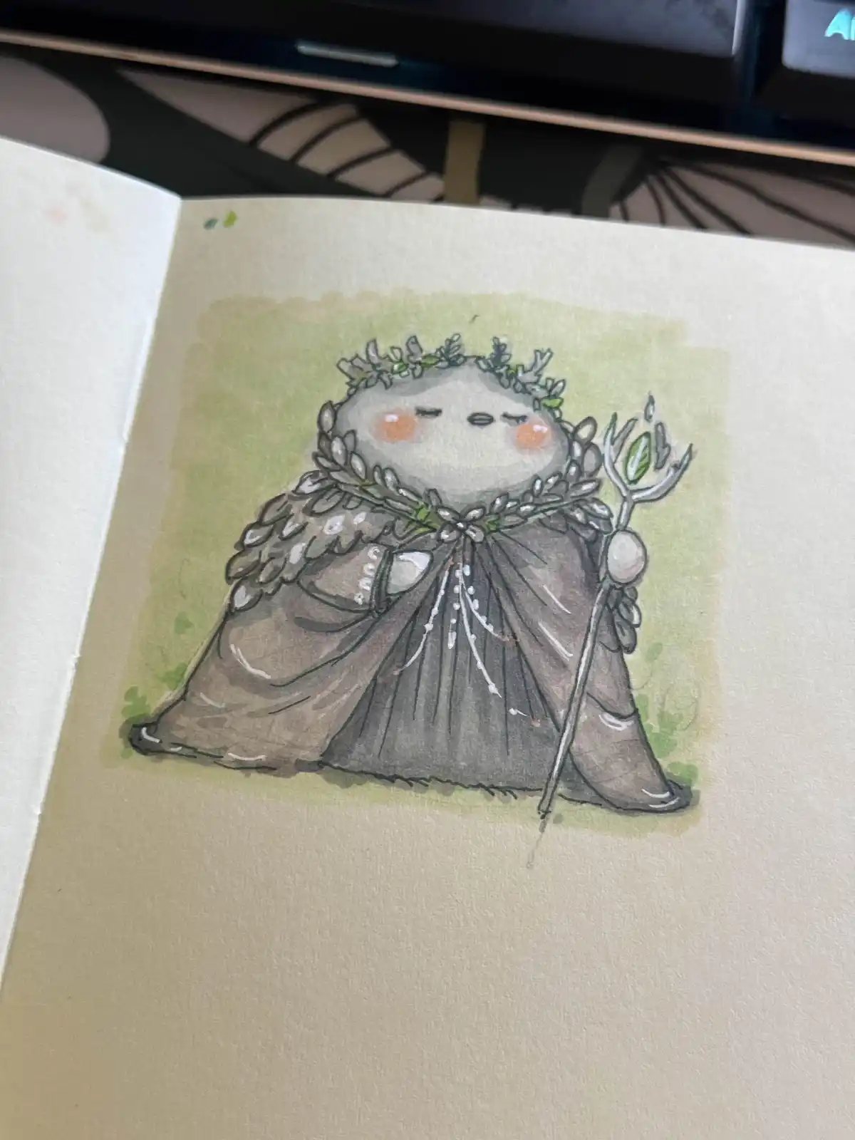 A watercolor drawing in a sketchbook shows a cute, round, grey creature with closed eyes and rosy cheeks, wearing a leafy…