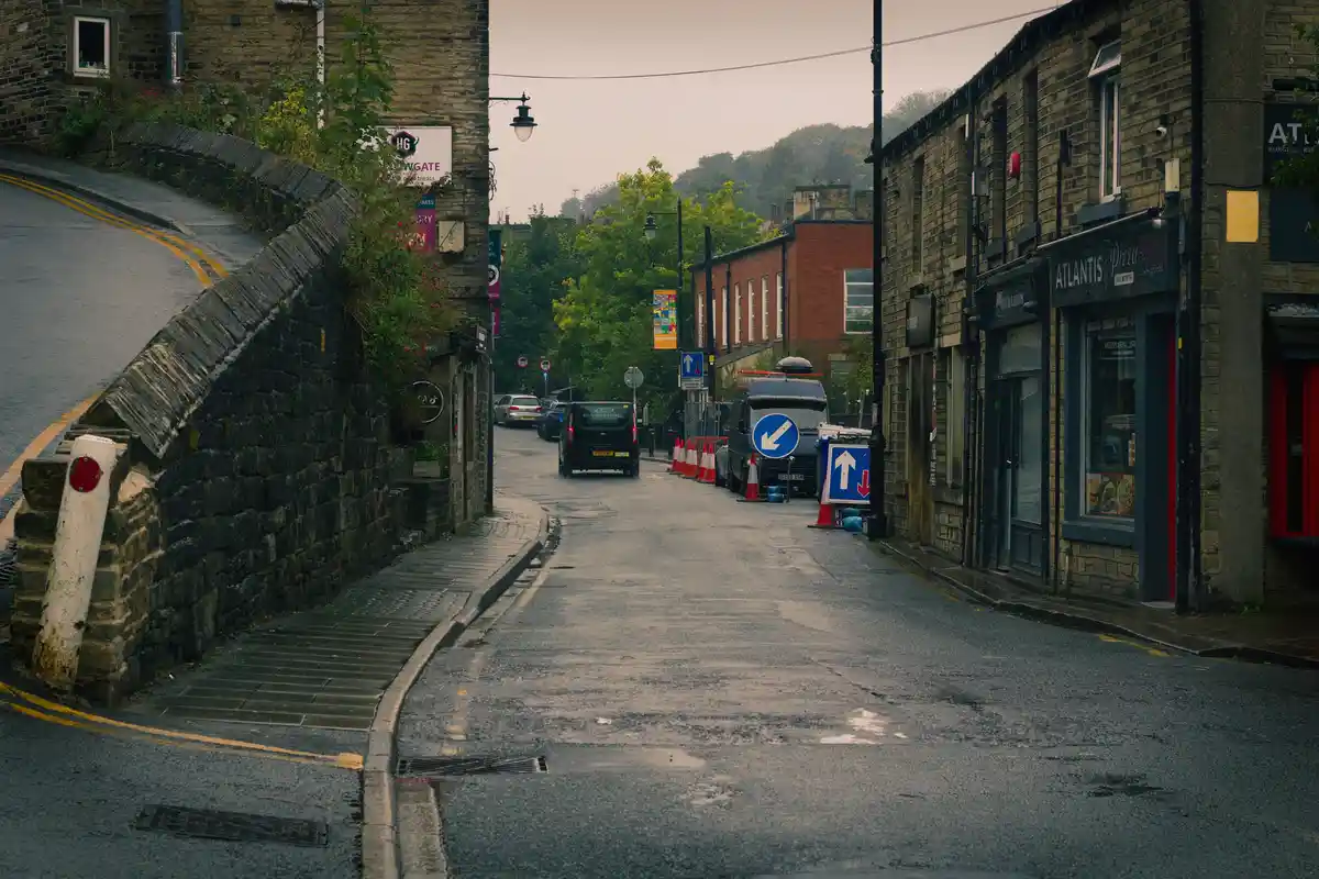 A wet, narrow street with stone walls and brick buildings on either side, featuring a black van and a white van parked near…