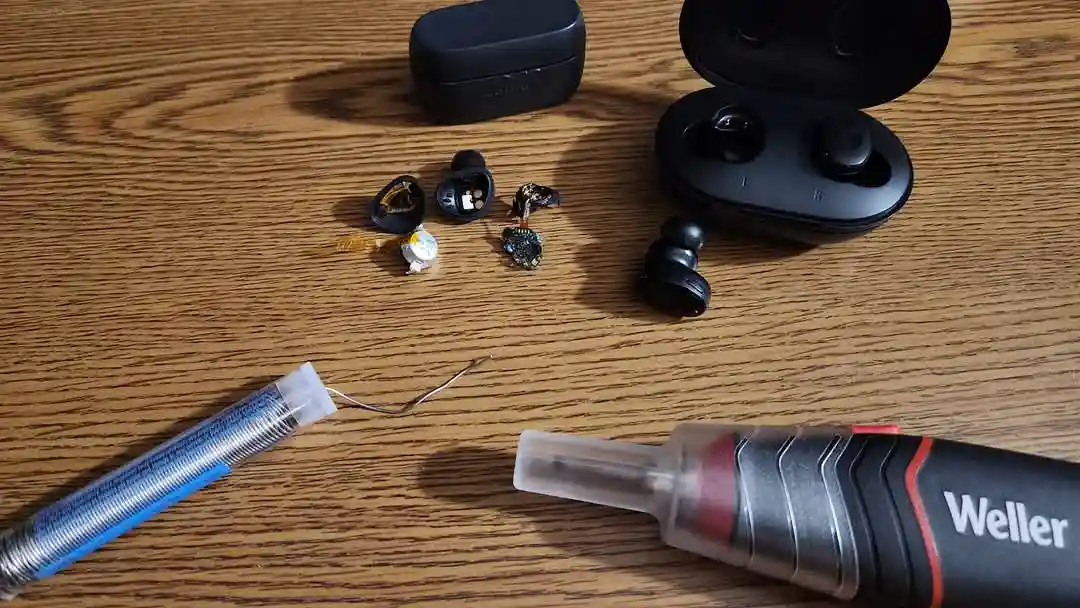 A wooden table displays a disassembled pair of black true wireless earbuds with their internal components scattered nearby…
