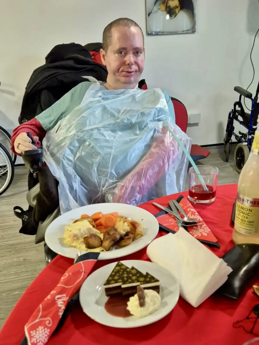 A man in a wheelchair, wearing a clear plastic bib, sits at a red tablecloth-covered table with a full Christmas dinner…