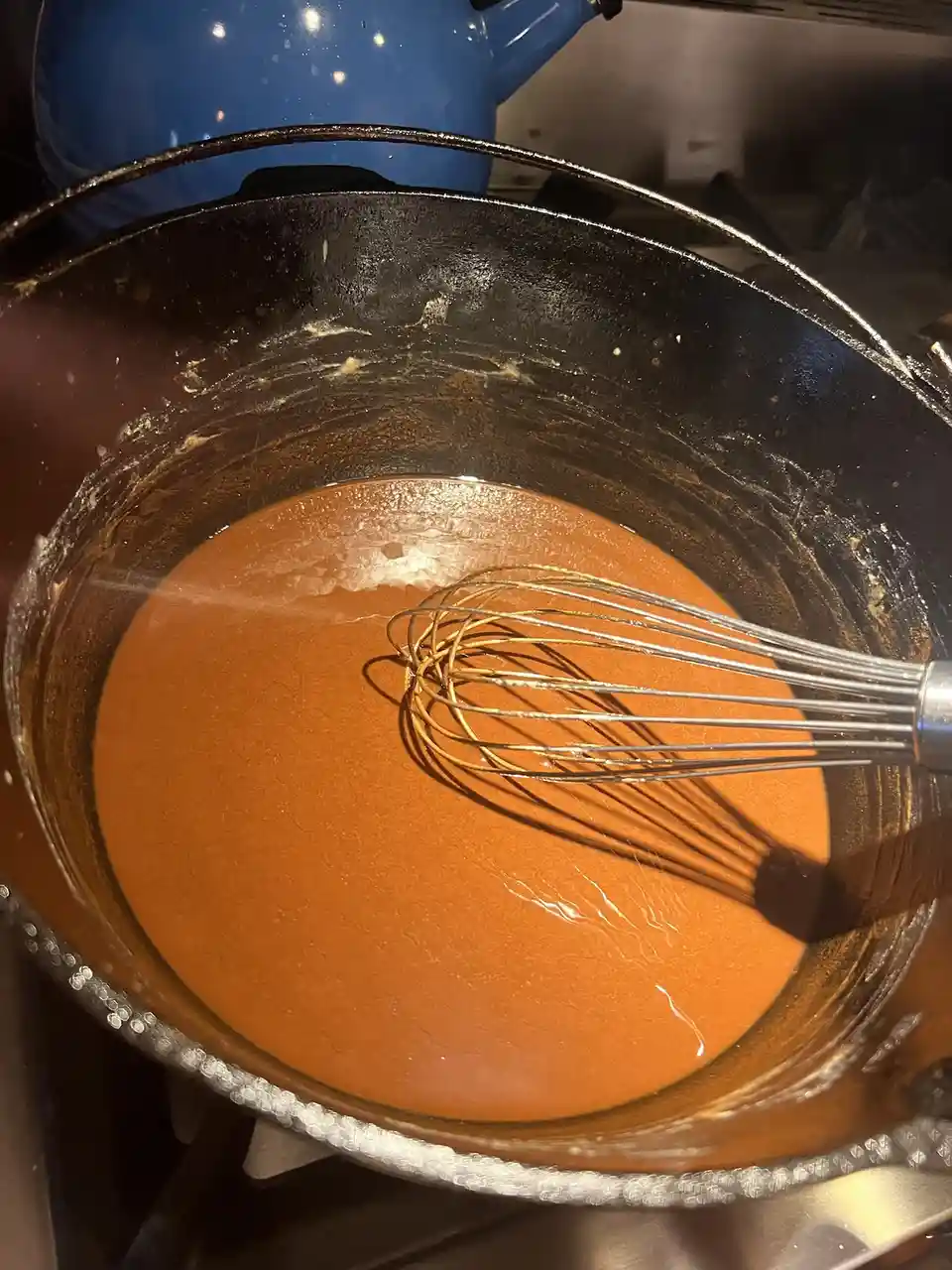 A close-up photo shows a dark, worn pot containing a smooth, orange-brown sauce with a metal whisk resting in it. In the…
