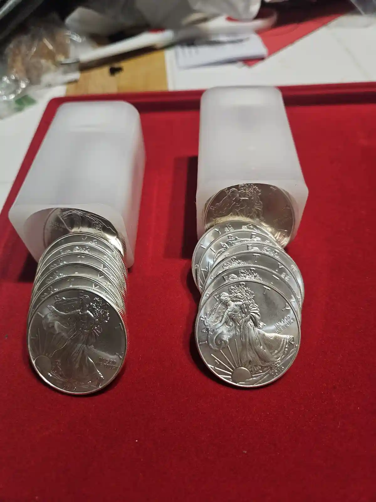Two stacks of silver American Eagle coins are shown spilling out of translucent plastic containers onto a red velvet…