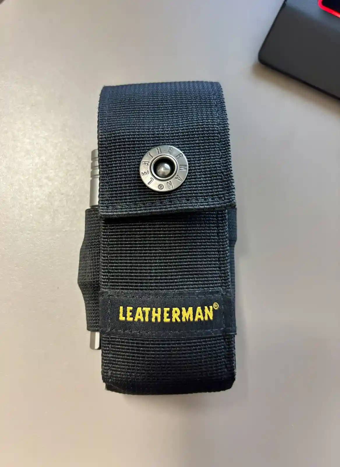 A black Leatherman multi-tool is partially visible inside a black fabric holster with a silver snap button and yellow…