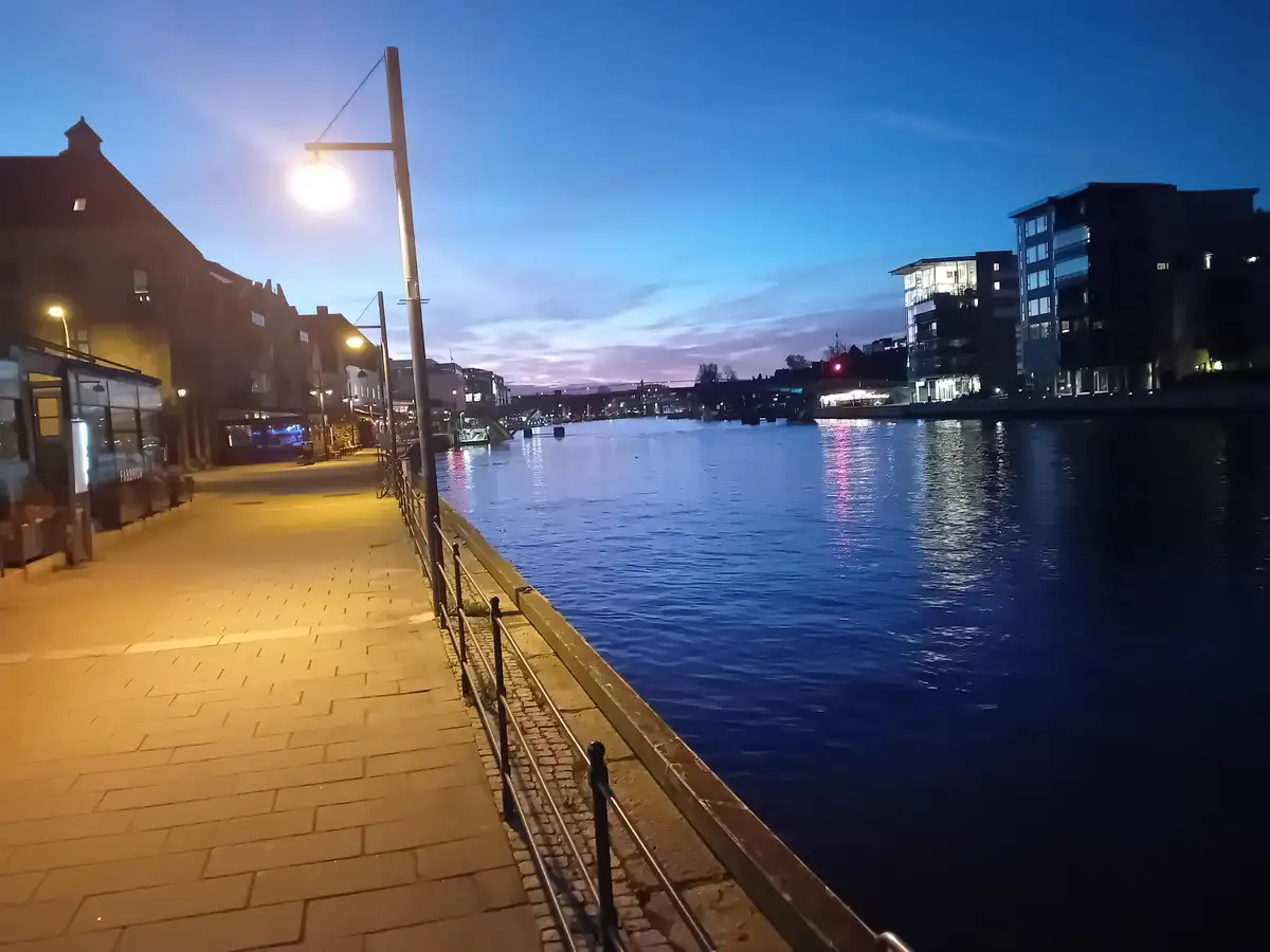 A serene twilight cityscape showing a paved riverside walkway with streetlights, a calm river reflecting city lights, and…
