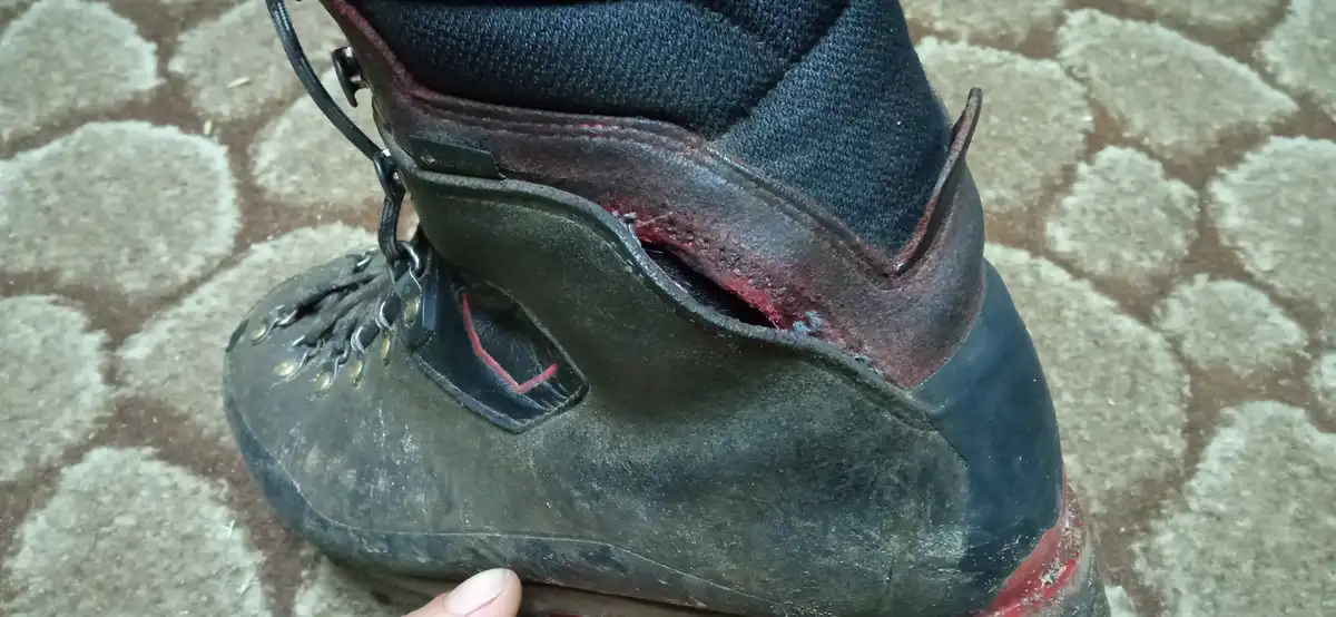 A close-up photo shows a heavily worn, dark-colored hiking boot with a large, torn opening at the ankle where red material…