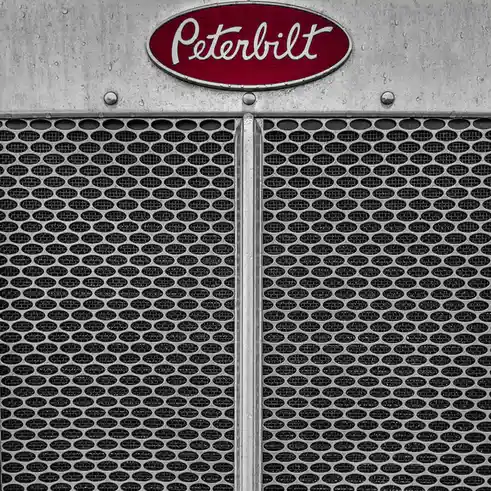 A close-up photo of a Peterbilt truck's front grille, featuring a red oval logo with white cursive text reading 'Peterbilt'…