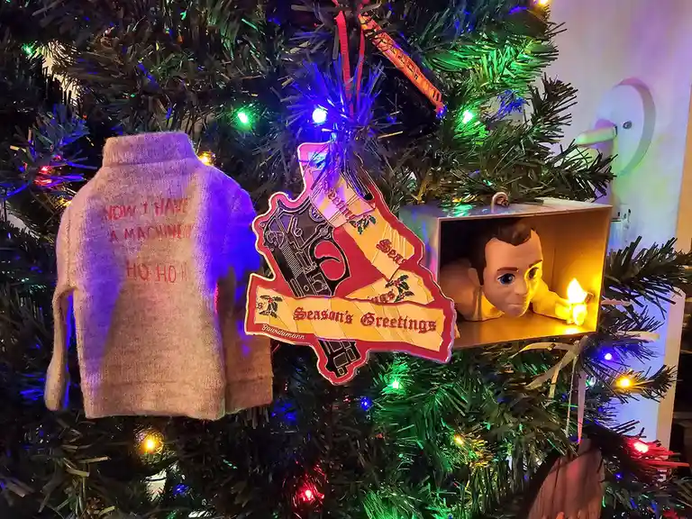 A close-up of a decorated Christmas tree with colorful lights, featuring three ornaments: a tiny sweater with red text…