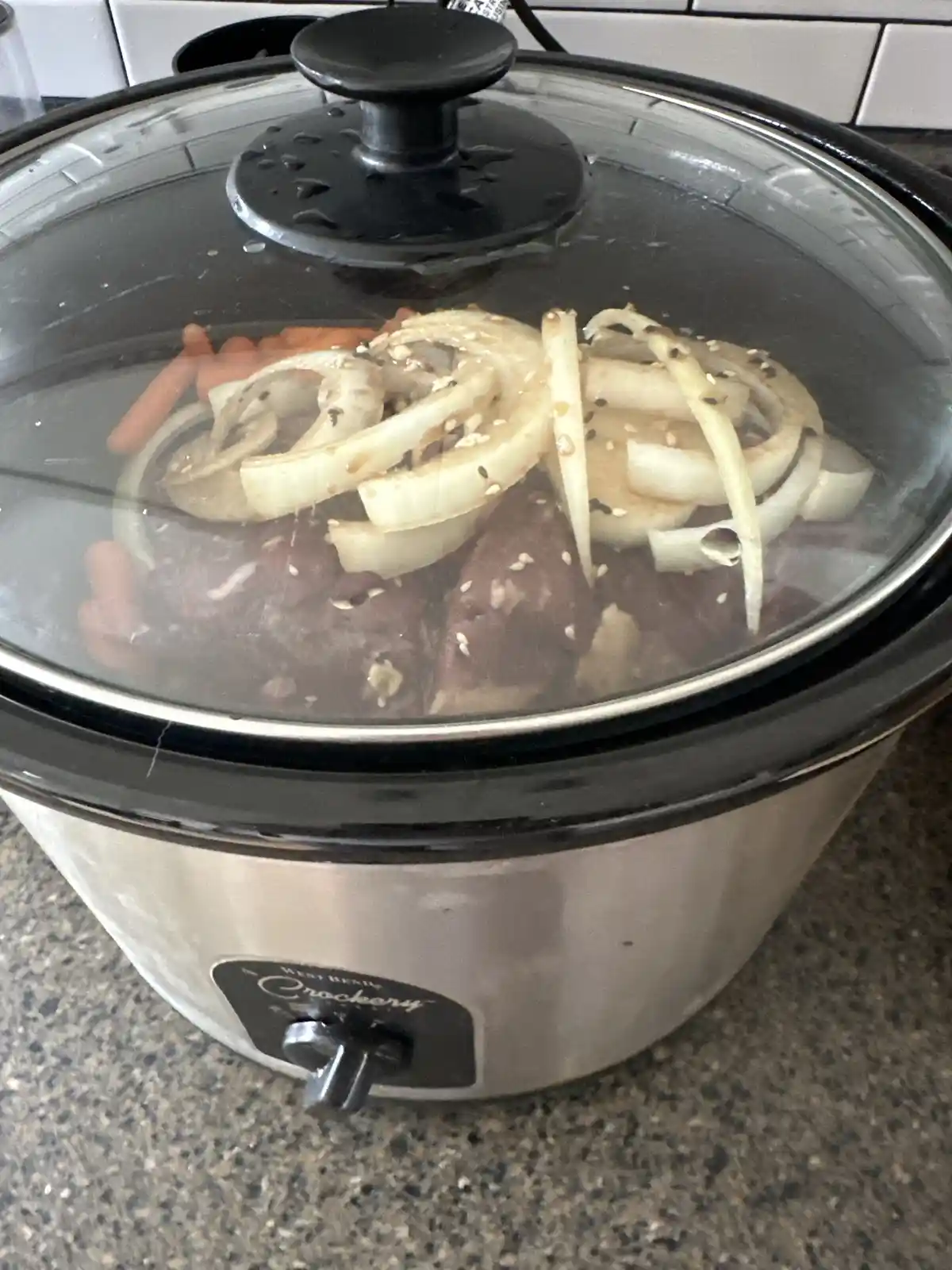 A stainless steel West Bend Crockery slow cooker with a glass lid sits on a countertop, showing cooked meat, sliced onions…