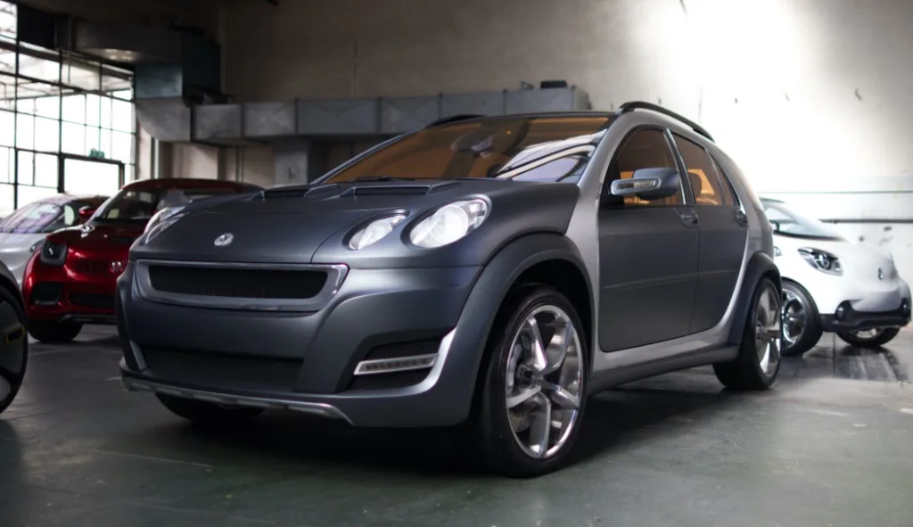 a dark gray compact car with a boxy, rugged design is prominently displayed in a showroom, flanked by other small cars…