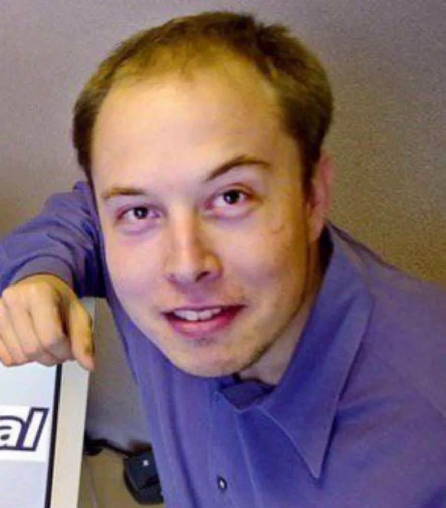 a close-up photo of a young man with thinning hair, wearing a purple collared shirt, leaning forward with his right hand on…