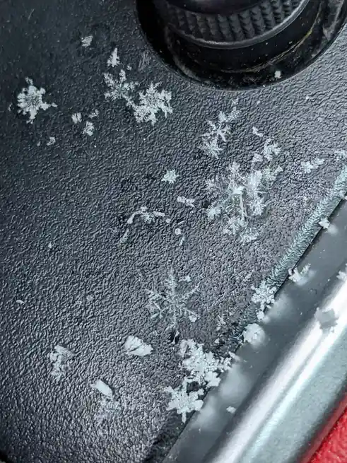 A close-up photo shows intricate, crystalline snowflakes resting on a dark, textured car interior surface, likely a seat or…
