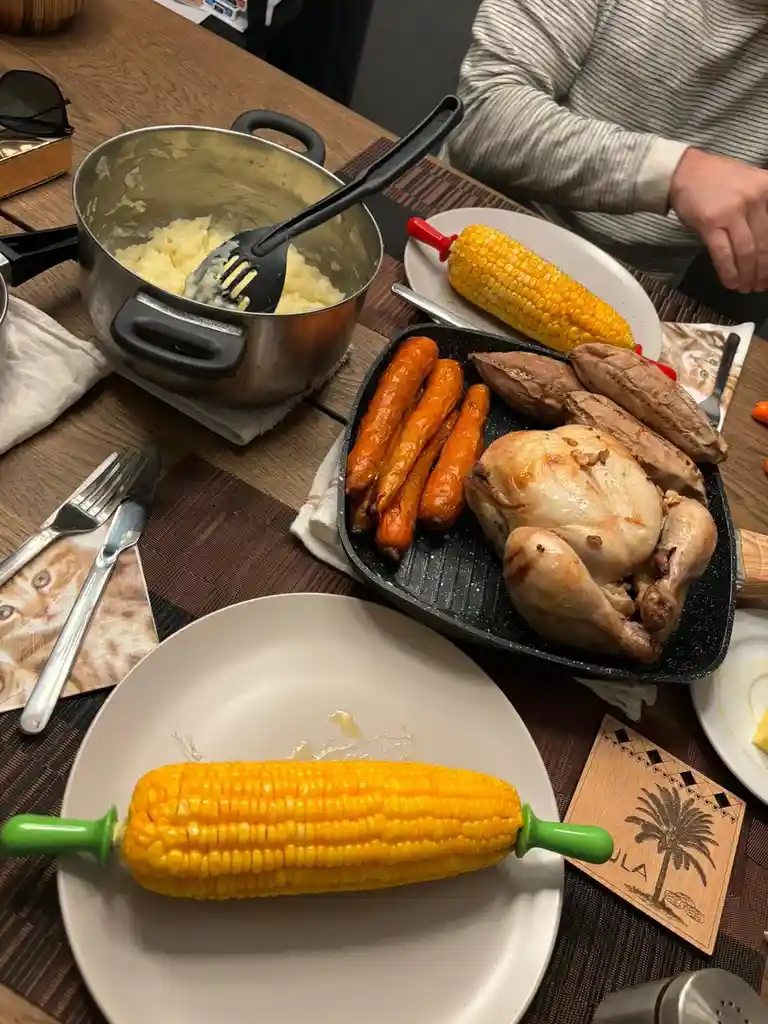 A table is set with a large roasted chicken, roasted carrots, sweet potatoes, and a pot of mashed potatoes, all appearing…