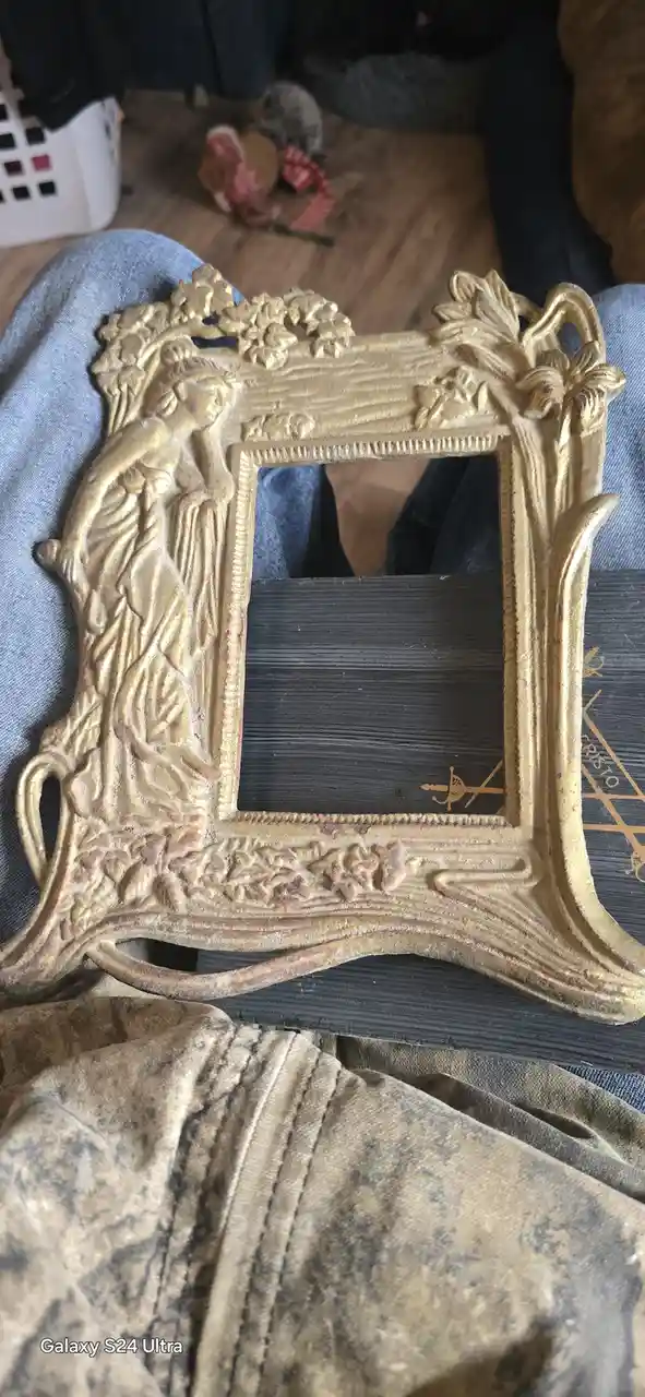 A close-up photo shows an ornate, gold-colored picture frame with intricate floral and figure carvings, resting on a…