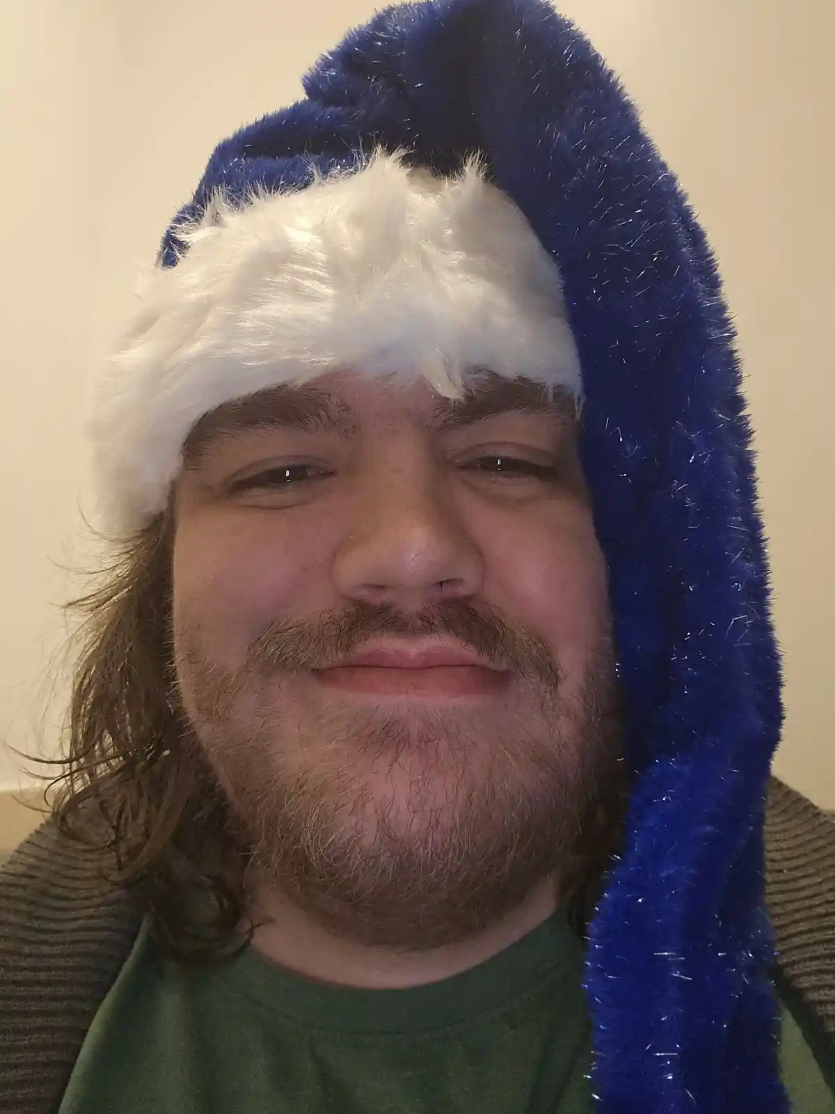 A man with a beard and mustache wears a blue and white fuzzy santa hat tilted to the side, looking at the camera with a…