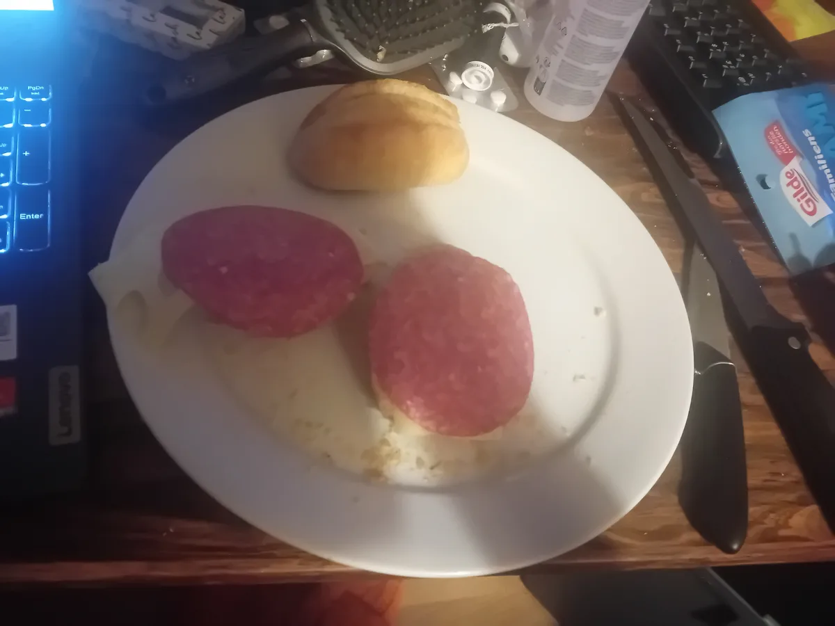 A white plate on a wooden desk holds a sandwich made with two round slices of salami, a slice of cheese, and a roll…