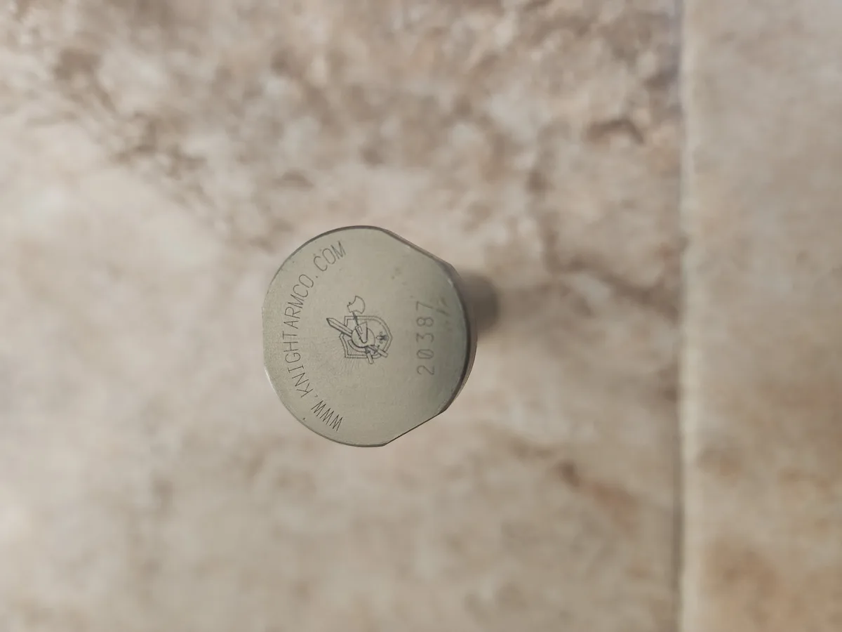 A close-up photo shows the top of a metal object, likely a tool or firearm part, resting on a beige marble-like surface. The…