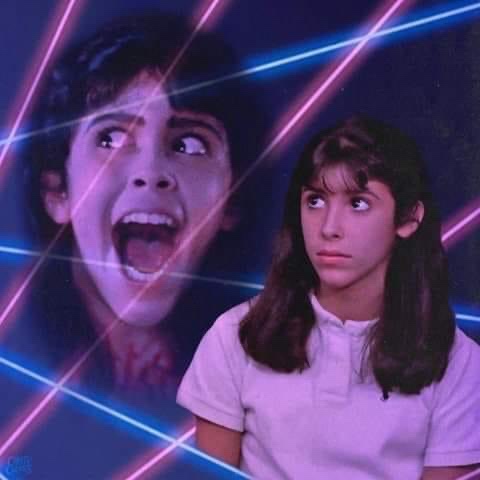 A still from the 1980s film 'The Lost Boys' shows a young woman in a white polo shirt looking calmly at the viewer, while a…