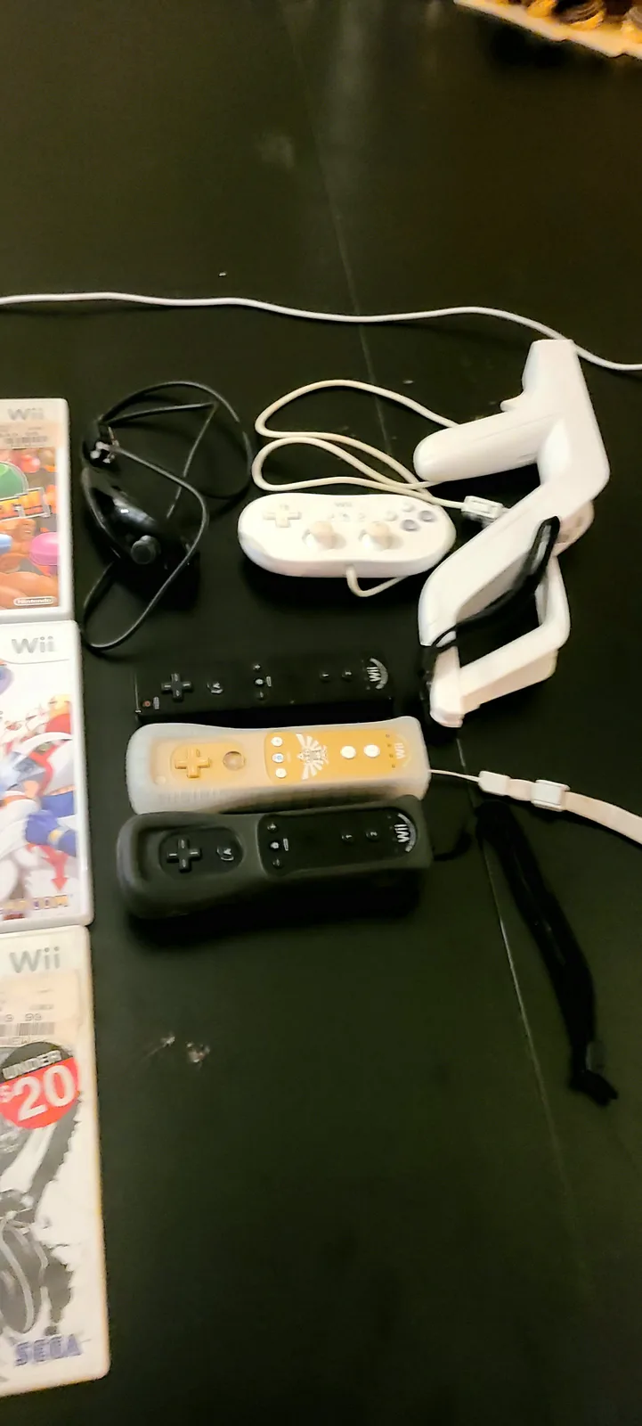 A collection of Nintendo Wii accessories, including multiple white and black Wii Remotes, a white Wii Wheel, a black…