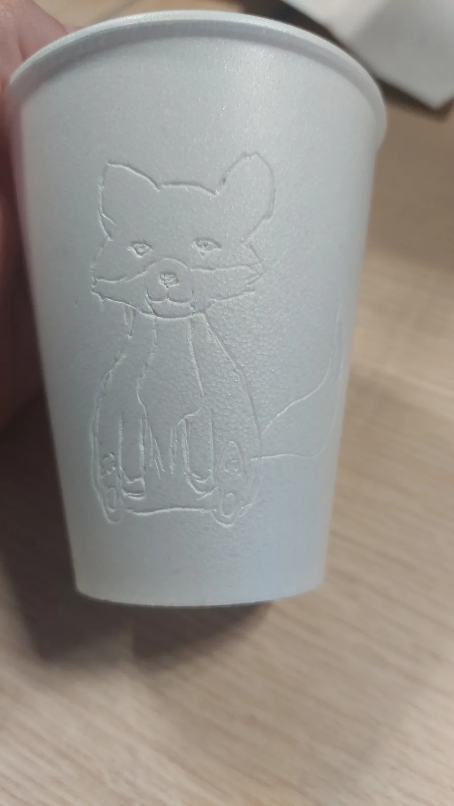 A close-up photo shows a white styrofoam cup with a raised, embossed image of a fox sitting upright on a scooter. The fox…