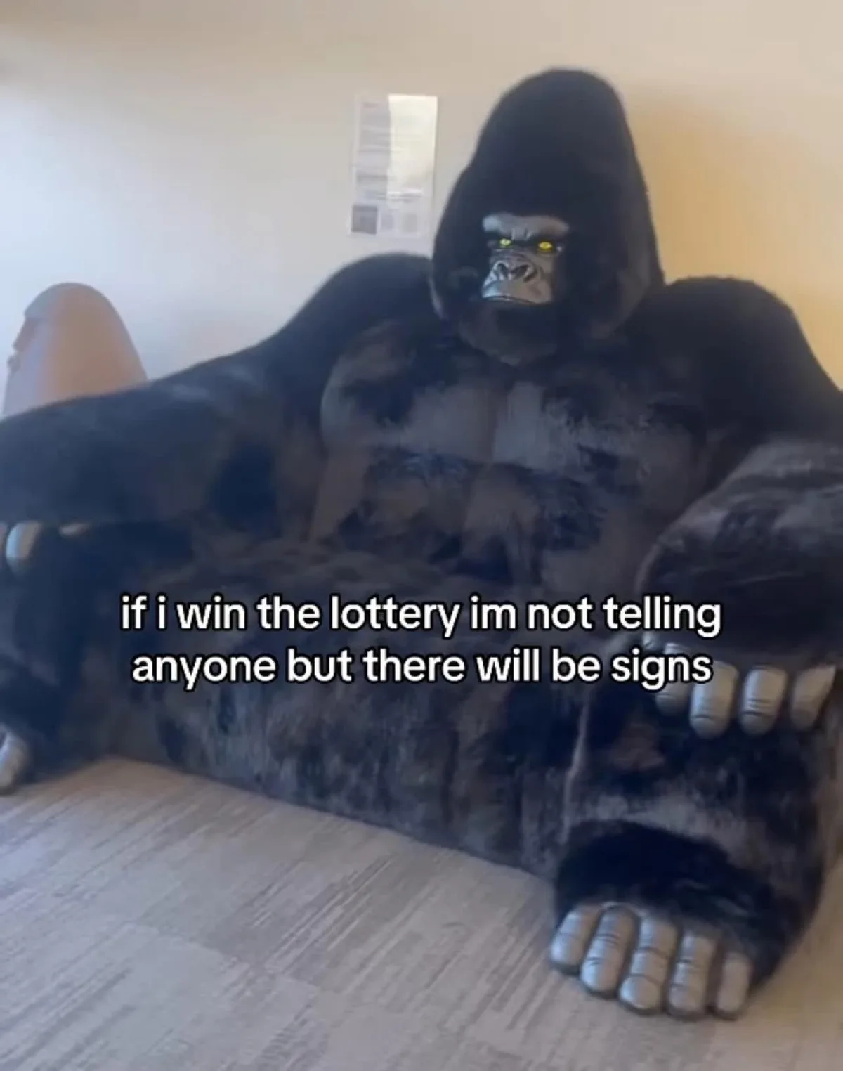 A large, black, furry gorilla costume is seated on a couch in a room, with glowing yellow eyes and plastic hands resting on…