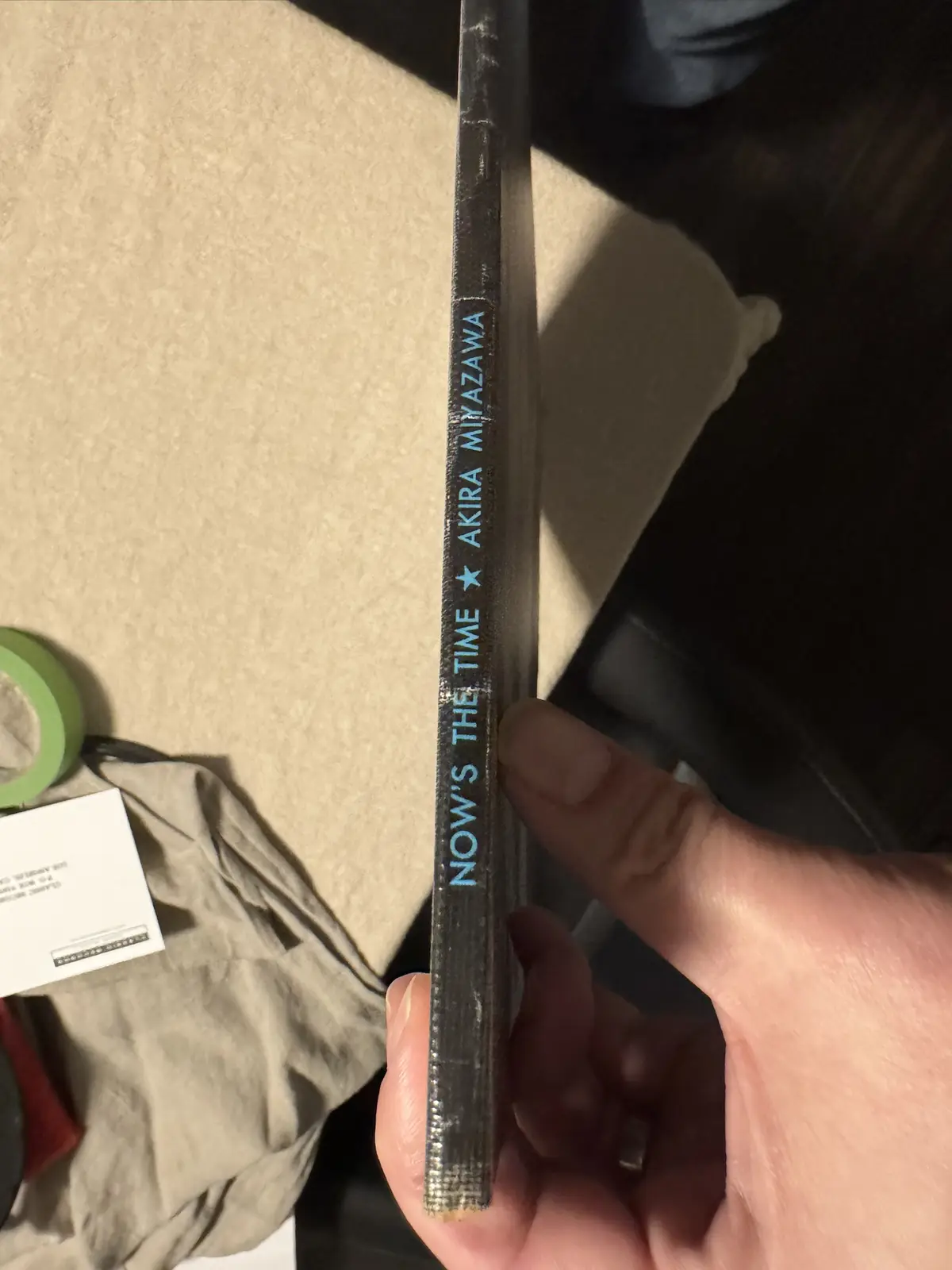 A hand holds a worn, dark-colored book spine vertically, showing the text 'NOW'S THE TIME ★ AKIRA MIYAZAWA' in light blue…