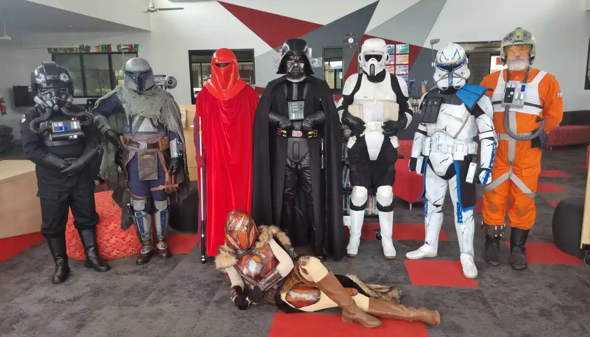 Seven people in Star Wars costumes stand in a line in a modern room with red and grey carpeting, while one person in a…
