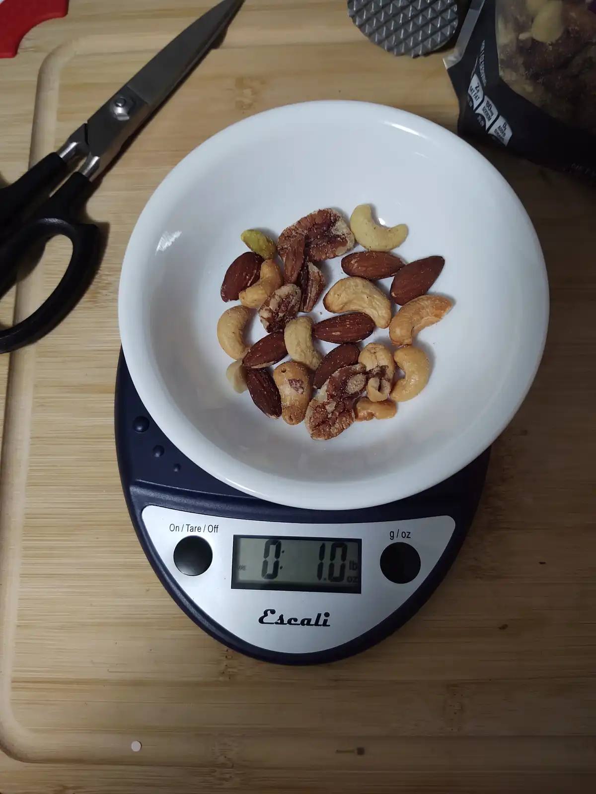 A white bowl filled with a mix of almonds, cashews, pecans, and pistachios sits on a digital kitchen scale that reads '0…