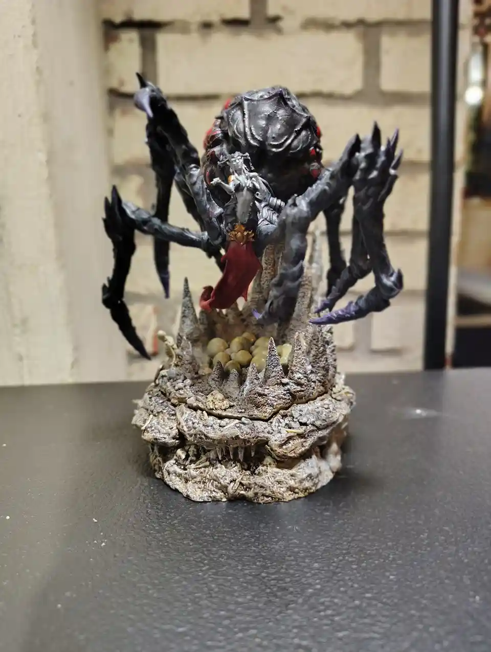 A detailed miniature figurine depicts a large, multi-limbed, black, monstrous creature with red accents, holding a small…