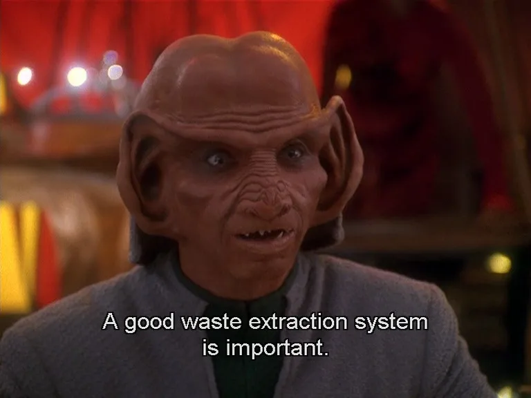 A close-up of a bald, grey-skinned alien with large ears and wrinkled forehead, wearing a grey uniform, looking shocked or…
