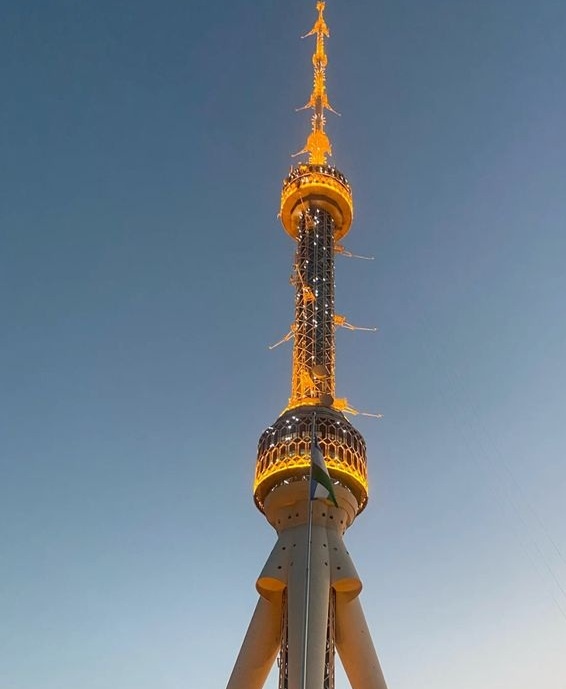 A low-angle photograph shows the top section of a tall, illuminated tower against a clear blue sky. The tower has a golden…