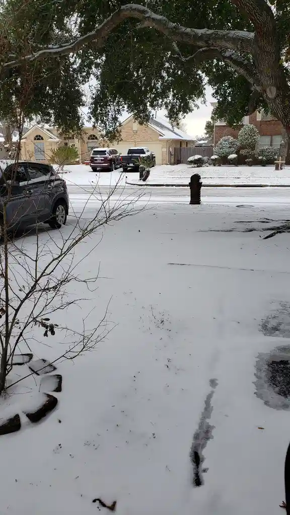 A suburban street in texas is covered in snow, with cars parked along the road and snow-dusted trees and houses visible in…