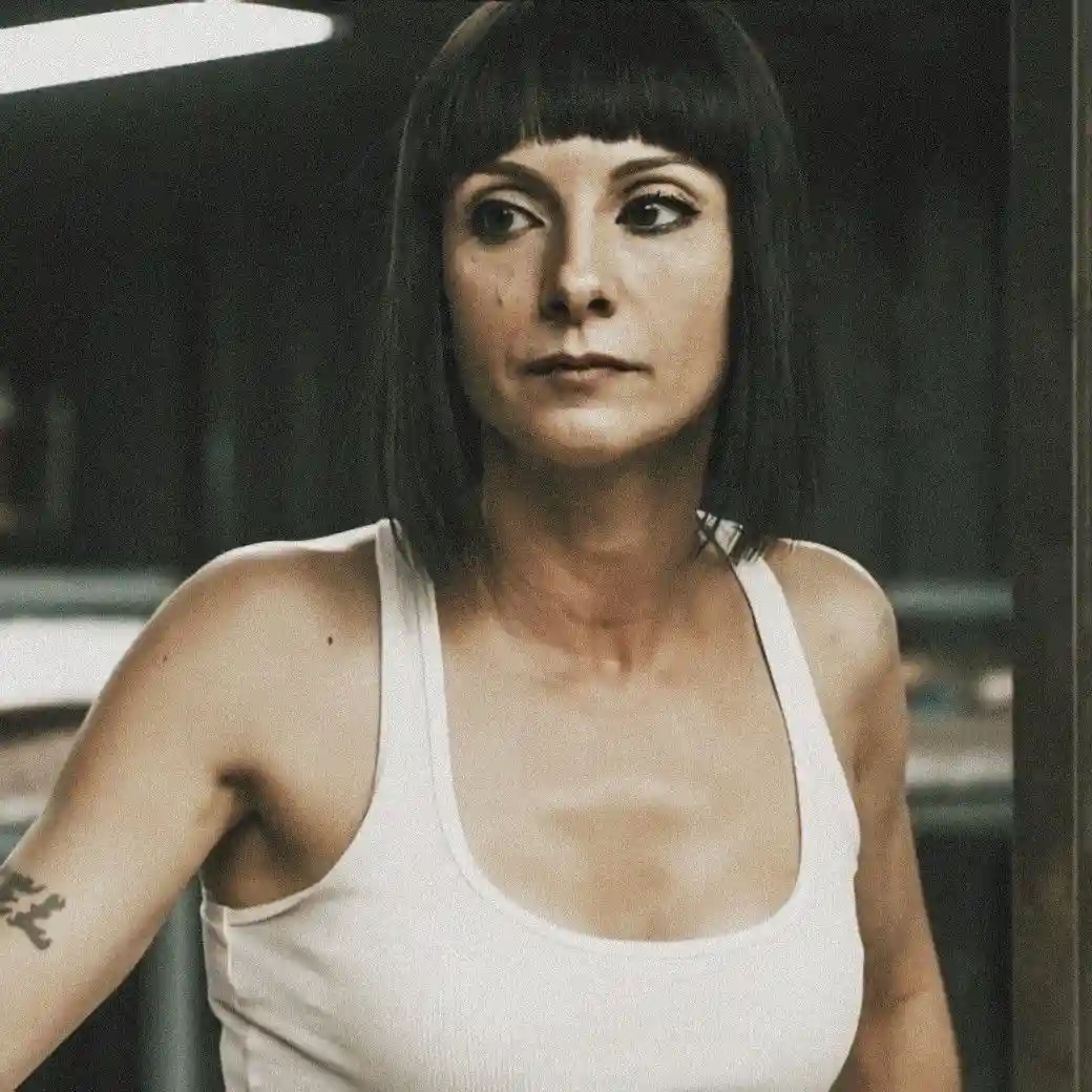 A woman with short black hair and bangs, wearing a white tank top, stares intently with a serious, unamused expression. She…