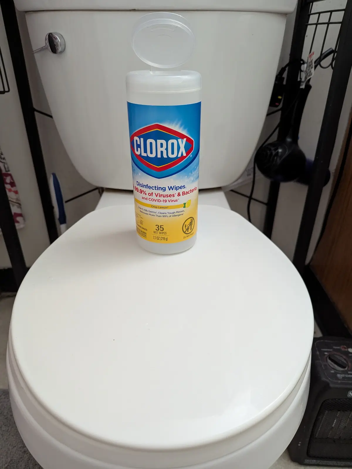 A bottle of Clorox disinfecting wipes sits on top of a closed white toilet lid, with the toilet tank visible behind it. The…