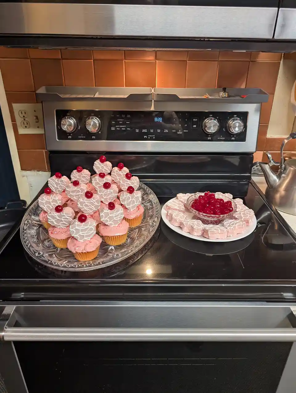A kitchen stove top displays two plates of pink-frosted desserts: cupcakes with white drizzle and cherry toppings on a glass…