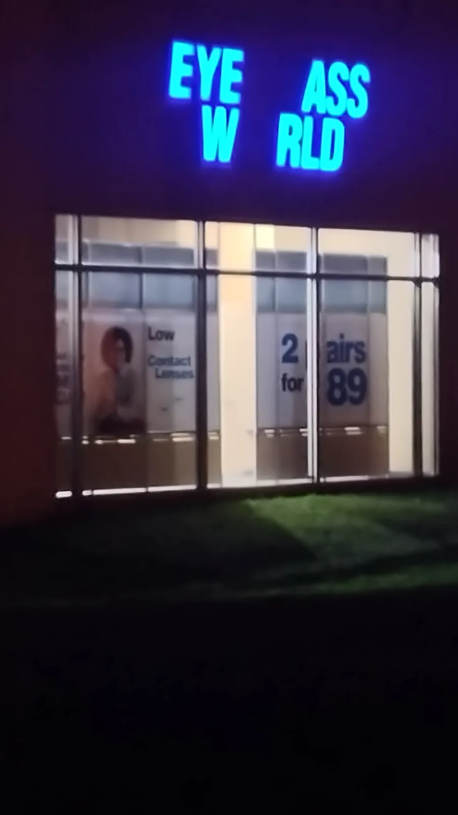 A nighttime photo of a store front with a blue neon sign that reads 'EYE W ASS RLD', which is a misspelling of 'Eye World'…