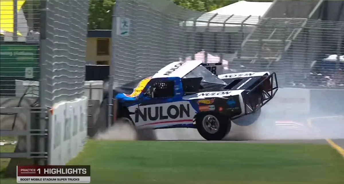 A blue, white, and black NULON-branded stadium super truck is airborne and drifting sideways, kicking up smoke as it crashes…