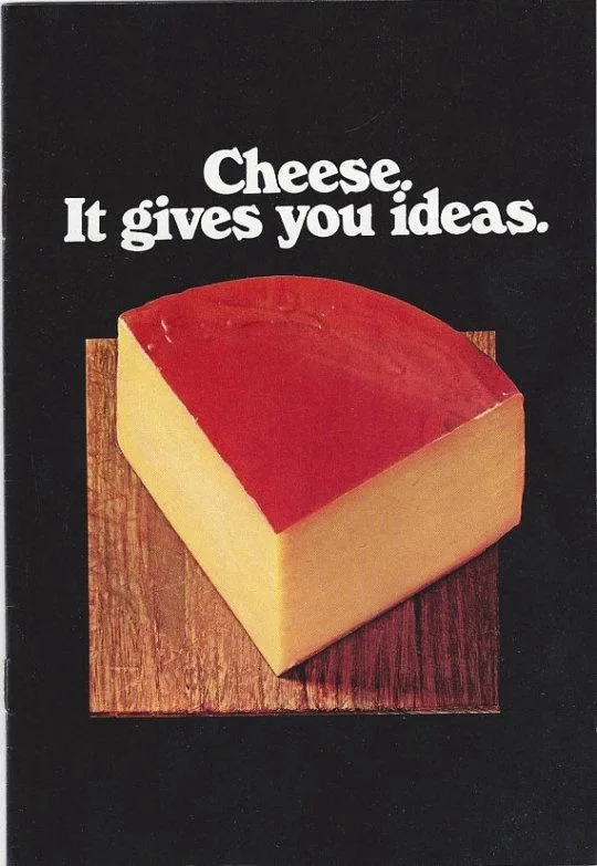 A vintage advertisement shows a large wedge of cheese with a red rind resting on a wooden board against a black background…
