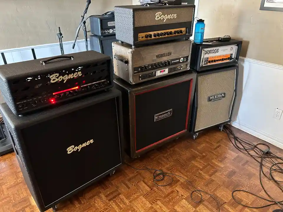 A room with several large, stacked guitar amplifiers from brands like Bogner, Verellen, and Mesa Engineering, sitting on a…