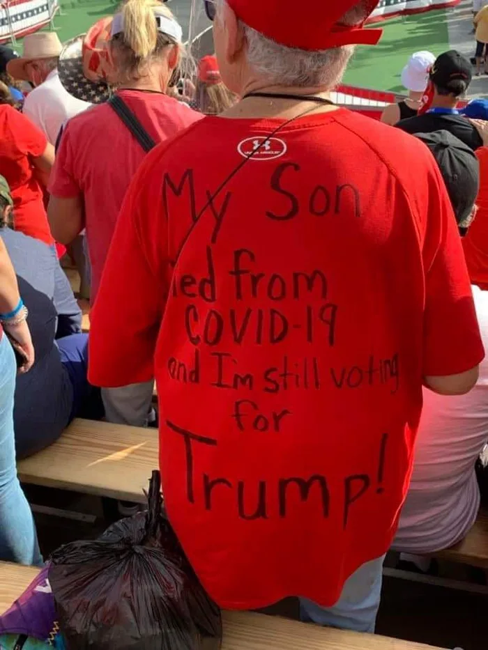A person wearing a red t-shirt with black handwritten text on the back that reads 'My Son died from COVID-19 and I'm still…