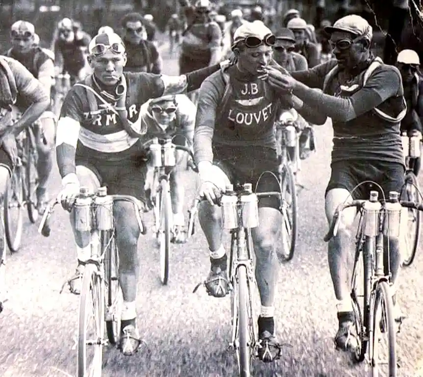 A vintage black-and-white photo shows three cyclists in a race, with one cyclist in the center wearing a jersey with 'J.B…