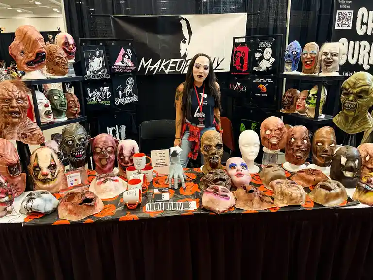 A woman wearing a white face mask stands behind a table covered in dozens of grotesque, horror-themed latex masks at a…